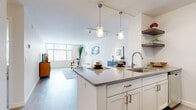 Located at 1254 Hennepin Ave N in Minneapolis, MN, this one-bedroom, one-bathroom residence offers a well-organized layout across a single floor. The open-concept living and kitchen area is bright and airy, featuring light hardwood-style flooring and large windows that frame sweeping views of the Minneapolis skyline. The kitchen is outfitted with white cabinetry, gray countertops, a stainless steel sink, subway tile backsplash, and stainless steel appliances including a range and over-the-range microwave. A peninsula with pendant lighting doubles as a dining and prep area, connecting seamlessly to the living room. The living room is generously proportioned, with large windows allowing abundant natural light and city views. The bedroom features plush carpeting, a sizeable window with urban views, and direct access to a walk-in closet equipped with wire shelving for organized storage. The bathroom includes wood-look tile flooring, white cabinetry with brushed nickel hardware, a tub-shower combination, and open wall shelving. A hallway connects the main living areas to the private bedroom wing, contributing to the unit's efficient and functional flow throughout.