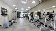This single-floor fitness center is thoughtfully designed to accommodate a wide range of workout styles and fitness goals. The main gym area features a patterned tile floor in a chevron layout, large wall-mounted mirrors, and a dropped grid ceiling with recessed LED panels and hanging pendant lights, creating a bright and motivating environment. Ceiling fans are distributed throughout to maintain airflow across the space.

The strength training section is well-appointed with a full rack of dumbbells, multiple cable and resistance machines, and dedicated benches. A climbing peg board and functional training wall add specialized options for advanced athletes. Large windows and glass doors allow natural light to flow in, offering views of the surrounding greenery.

The cardio zone lines one wall with a row of treadmills and ellipticals positioned in front of arched windows, enhancing the workout experience with natural light and outdoor views. Soaring vaulted ceilings distinguish this section of the facility.

A dedicated turf training area features a marked indoor turf surface with yard lines, a RAX functional training rig stocked with resistance bands, medicine balls, kettlebells, a BOSU ball, a battle rope, and weight plates — ideal for high-intensity and functional fitness training. A spin bike and open floor space complete this versatile zone, supporting everything from individual conditioning to group exercise sessions.