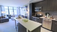 This single-floor residence showcases a thoughtfully designed open-concept layout that integrates the kitchen and living areas into one cohesive, functional space. The kitchen is a standout feature, equipped with stainless steel appliances including a range with oven, over-the-range microwave, and dishwasher. Dark espresso cabinetry extends from floor to ceiling, providing ample storage, while a mosaic tile backsplash adds a refined textural detail. The light-toned countertops offer generous workspace and extend to a peninsula-style island with seating, making it ideal for casual dining or entertaining. The island is finished with a sleek quartz-style surface and a barstool tucked neatly underneath. Floating shelves with decorative accents add visual interest along the wall adjacent to the living area. The living space benefits from large floor-to-ceiling windows that invite abundant natural light and offer views of surrounding greenery. The overall ambiance is contemporary and clean, with a neutral color palette punctuated by dark cabinetry contrasts, creating a polished and inviting atmosphere throughout.