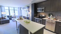 This single-floor residence showcases a thoughtfully designed open-concept layout that integrates the kitchen and living areas into one cohesive, functional space. The kitchen is a standout feature, equipped with stainless steel appliances including a range with oven, over-the-range microwave, and dishwasher. Dark espresso cabinetry extends from floor to ceiling, providing ample storage, while a mosaic tile backsplash adds a refined textural detail. The light-toned countertops offer generous workspace and extend to a peninsula-style island with seating, making it ideal for casual dining or entertaining. The island is finished with a sleek quartz-style surface and a barstool tucked neatly underneath. Floating shelves with decorative accents add visual interest along the wall adjacent to the living area. The living space benefits from large floor-to-ceiling windows that invite abundant natural light and offer views of surrounding greenery. The overall ambiance is contemporary and clean, with a neutral color palette punctuated by dark cabinetry contrasts, creating a polished and inviting atmosphere throughout.