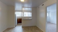 This single-floor space features a straightforward, functional layout with a notable open pass-through design connecting the kitchen and living area. The kitchen is equipped with dark wood cabinetry, a sink positioned beneath a bright window that allows natural light to filter in, and a white refrigerator. The pass-through window between the kitchen and the adjacent living space enhances the sense of openness and facilitates easy interaction between the two areas. The flooring throughout is a warm-toned tile that provides a cohesive look across the space. Walls are painted in a clean, neutral white, creating a bright and airy atmosphere that complements the natural light coming through the kitchen windows. Ceiling-mounted light fixtures provide additional illumination. The living area offers a blank canvas with ample wall space, making it adaptable for various furniture arrangements. An entry door with a brass-toned handle leads into the space, with a light switch conveniently positioned nearby. The overall design is practical and straightforward, making efficient use of the available floor plan.