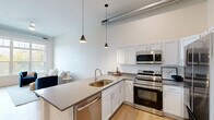 Located at 1107 S 44th St in Cumming, Iowa, this modern one-bedroom, one-bathroom unit offers a well-designed, single-floor layout that maximizes both functionality and style. The open-concept kitchen and living area serves as the heart of the unit, featuring white shaker cabinetry, gray quartz-style countertops, a peninsula with an undermount sink, and a full suite of stainless steel appliances including a range, over-the-range microwave, and dishwasher. Sleek pendant lighting and exposed industrial ductwork overhead add a contemporary loft-inspired character, while light hardwood flooring flows throughout the space. The living area benefits from large windows that draw in abundant natural light and offer views of the surrounding landscape. The bedroom is a comfortable retreat with soaring ceilings, an exposed duct detail, and a tall window providing natural light and outdoor views. It connects directly to a walk-in closet outfitted with wire shelving. The full bathroom features a clean, white palette with a vanity, tub/shower combo, and hardwood flooring. A dedicated laundry room with a side-by-side washer and dryer completes this thoughtfully appointed unit.