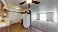 This single-floor unit offers a practical, open layout that seamlessly connects the kitchen and living area. The kitchen is equipped with white appliances, including a refrigerator, electric range, and microwave, complemented by dark wood cabinetry mounted above for additional storage. Hardwood-style flooring runs through the kitchen, providing a clean, durable surface that transitions into carpeting in the adjoining living area. A ceiling fan with a light fixture is centrally positioned overhead, enhancing air circulation and illumination throughout the shared space. The textured ceilings add a subtle visual detail consistent throughout the unit. The living area is notably bright, benefiting from a large sliding glass door fitted with vertical blinds, which allows generous natural light to fill the space and provides access to the outdoors. The overall layout is compact yet functional, with a clear division between the kitchen and living zones created by a partial wall. The neutral color palette, combining white walls and warm wood tones, creates a straightforward and welcoming ambiance suited to everyday living.