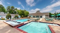 Located at 9440 Pleasant Point Way in Ashland, Virginia, this outdoor recreational facility offers a well-appointed aquatic complex designed for relaxation and leisure. The centerpiece is a generously sized swimming pool with clear blue water, framed by brick-accented edges and dark blue tile detailing, providing a classic and polished aesthetic. A separate spa or wading pool area is positioned adjacent to the main pool, offering a more intimate retreat. The expansive sundeck surrounding the pools is outfitted with numerous lounge chairs and teal-accented umbrellas, providing ample space for sunbathing and socializing. A white pergola structure on one side of the deck offers a shaded gathering area with additional seating. The single-story clubhouse building, featuring a distinctive cupola roofline, a pitched shingle roof, and large glass doors, anchors one end of the complex and opens directly onto an elevated deck overlooking the pool area. The entire complex is enveloped by mature trees, creating a private, park-like atmosphere. The well-maintained landscaping, including manicured shrubs and seasonal plantings, enhances the overall visual appeal. The layout efficiently balances open sunlit areas with shaded retreats, making this a versatile outdoor amenity suitable for a wide range of recreational activities throughout the warmer months.