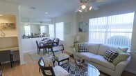 This single-floor residence showcases an open-concept layout that seamlessly connects the living and dining areas, creating a spacious and cohesive environment. The living area is anchored by a plush, neutral-toned sofa accented with patterned throw pillows, complemented by a glass-top coffee table and stylish accent chairs upholstered in a bold animal print. A ceiling fan with integrated lighting fixture hangs overhead, providing both function and a finishing touch to the space.

Large windows dressed with vertical blinds line the walls, allowing generous amounts of natural light to flood the interior, enhancing the warm tones of the light hardwood flooring throughout. Decorative artwork adorns the walls, adding color and personality to the neutral palette.

The adjacent dining area accommodates a full dining set with dark wood chairs and a matching table, set atop an area rug. A mirrored wall feature in the dining space adds depth and amplifies the sense of openness. The overall ambiance is bright, modern, and welcoming, with cohesive décor choices that tie the connected spaces together harmoniously.