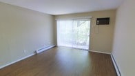 This single-floor space is designed with functionality and comfort in mind. The main living area showcases warm hardwood-style flooring that extends throughout, lending a cohesive and inviting feel to the room. A large sliding glass door serves as the focal point of the space, fitted with vertical blinds that allow for flexible light control. The door provides a view of mature greenery outside, bringing a natural element into the interior and filling the room with abundant natural light when the blinds are open.

Climate control is well-covered, with a baseboard heating unit running along the exterior wall beneath the sliding door, ensuring efficient warmth during cooler months. A wall-mounted air conditioning unit on the adjacent wall provides cooling during warmer seasons, making the space comfortable year-round.

The walls are finished in a neutral beige tone, complementing the flooring and contributing to a clean, understated aesthetic. The open layout of the living area offers flexibility for a variety of furniture arrangements. Overall, the space presents a practical and straightforward design with key functional features thoughtfully incorporated.