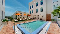 Located at 125 Quadrille Plaza Dr in the heart of West Palm Beach, Florida, this outdoor amenity space offers a well-appointed pool deck set within a multi-story building courtyard. The centerpiece is a sparkling rectangular swimming pool finished with vivid blue mosaic tiles, equipped with stainless steel entry rails and a shallow wading entry — ideal for a range of guests. The pool deck is surfaced in warm terracotta-toned pavers that complement the crisp white stucco façades of the surrounding building. A dedicated lounge area features white chaise loungers shaded by bold orange market umbrellas, arranged on a patch of artificial turf for a clean, resort-like aesthetic. Tropical plantings, including palms and lush greenery, are thoughtfully integrated into raised planters along the perimeter, adding a vibrant natural element to the urban setting. A walkway with a railed balcony overlooks the surrounding West Palm Beach streetscape, providing open views of the city's architectural character. The open-air design allows for abundant natural sunlight throughout the day, creating an inviting and relaxing outdoor retreat. The space is directly accessible from the building's interior, making it a convenient and attractive amenity for occupants.