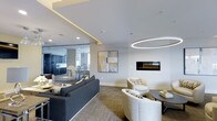 This thoughtfully designed single-floor space offers a seamless open-concept layout that connects a refined lounge area, a modern kitchen, and a dining space. The lounge is anchored by a sleek, wall-mounted linear fireplace that serves as a striking focal point, complemented by a large-scale abstract artwork in muted gold and grey tones. A bold circular pendant light fixture adds a dramatic architectural element overhead, while recessed lighting and an integrated cove light detail along the ceiling contribute to a warm, layered ambiance. The seating arrangement features a plush charcoal sofa paired with curved ivory accent chairs, creating an inviting and contemporary gathering area. Underfoot, a textured carpet in neutral tones grounds the space with subtle warmth. A full-height glass partition elegantly separates the lounge from the kitchen and dining area beyond, maintaining visual connectivity while defining distinct functional zones. The kitchen is outfitted with dark cabinetry, modern appliances, and pendant lighting above a dining table, conveying a refined and well-appointed culinary space. Throughout, the design palette of crisp whites, deep charcoals, and gold accents delivers a cohesive, upscale aesthetic.