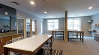 This single-floor communal space is thoughtfully designed to accommodate a variety of uses, from group work sessions to casual socializing. The room features a neutral color palette of soft grays and warm wood tones, creating a welcoming and professional atmosphere throughout.

At the forefront is a large, light-colored stone-top table paired with modern wire-frame chairs, providing an ideal surface for collaborative tasks or shared meals. Beyond it, two additional high-top tables with matching seating are arranged near the perimeter, offering alternative spots for smaller groups.

Large windows line the far wall, allowing generous amounts of natural light to fill the space and providing views of the surrounding exterior. The carpeted flooring, featuring a subtle striped pattern near the main table, adds a layer of warmth underfoot.

A structural column with a wood-finished base anchors the center of the room without interrupting its open, airy feel. To the left, glass-paneled doors lead to what appears to be a separate enclosed room or office area, adding functional versatility to the overall layout. Recessed ceiling lighting supplements the natural light, ensuring the space remains well-lit throughout the day.
