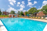 This outdoor pool area offers a well-appointed recreational space designed for warm-weather enjoyment. The centerpiece is a generously sized swimming pool with clear, blue-green water, accessible via built-in entry steps and a metal ladder. The surrounding concrete sundeck provides ample room for relaxation and circulation, lined with a row of orange lounge chairs and shaded by multiple teal market umbrellas, including a dedicated table-and-chair seating area ideal for socializing.

A single-story brick pool house serves as a functional anchor to the space, offering storage for safety equipment such as life jackets and rescue gear. A prominently posted sign notes that no lifeguard is on duty and that children must be supervised at all times. The pool house features a window overlooking the pool area and is accessed via a standard entry door.

The entire pool area is enclosed by a black wrought iron fence, providing both security and an open, airy feel. Lush mature trees and manicured green lawns frame the perimeter, offering natural shade and a pleasant backdrop. Brick residential buildings are visible beyond the fence, indicating this is a shared community amenity. The space is bathed in abundant natural sunlight, creating a bright and inviting outdoor atmosphere.
