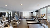 Located at 55 Melrose Terrace in Long Branch, NJ, this fitness center is a fully outfitted exercise facility designed to support a comprehensive workout routine. The single-floor space is finished with durable rubber flooring suited for high-traffic use, and the smooth white ceiling is fitted with recessed lighting that ensures bright, even illumination throughout. Large windows line the walls, inviting generous natural light and offering views of the surrounding greenery, creating an open and energizing atmosphere. Full-length mirrors are mounted along one side of the room, visually expanding the space and allowing users to monitor their form during exercise. The cardio section is well-stocked with multiple treadmills, elliptical machines, and stationary bikes from brands including TRUE. Strength training is accommodated by a multi-station cable machine, a dumbbell rack with a range of free weights, and additional resistance equipment. A wall-mounted television is positioned for easy viewing during cardio sessions. Access to the space is provided through glass-paneled double doors, and a hand sanitizer station is conveniently placed near the entrance. The overall design is clean and functional, with a neutral gray color palette that contributes to a focused, professional fitness environment.
