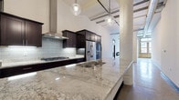 Located in Milwaukee's Historic Third Ward, this single-floor loft at 226 N Water St masterfully combines industrial heritage with contemporary design. Soaring ceilings, exposed concrete columns, polished concrete floors, and original brick walls set a striking architectural tone throughout. The open-concept kitchen is a standout feature, equipped with dark espresso cabinetry, a gas cooktop with a stainless steel range hood, stainless steel appliances, a subway tile backsplash, and an expansive granite island with an undermount sink — all illuminated by globe pendant lighting. The living area offers a sleek, wall-mounted linear fireplace and direct access to additional rooms via bold black interior doors. Exposed brick and large windows invite natural light while framing views of the surrounding Milwaukee streetscape. A dedicated laundry room provides in-unit washer and dryer hookups with additional cabinetry. The bathroom features white shaker cabinetry paired with dark granite countertops and an exposed brick accent wall. A generously sized walk-in closet includes a full wall of built-in shelving and drawers, offering substantial storage. Visible ductwork and industrial-style details throughout lend an authentic loft aesthetic to this refined urban residence.
