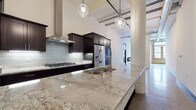 Located in Milwaukee's Historic Third Ward, this single-floor loft at 226 N Water St masterfully combines industrial heritage with contemporary design. Soaring ceilings, exposed concrete columns, polished concrete floors, and original brick walls set a striking architectural tone throughout. The open-concept kitchen is a standout feature, equipped with dark espresso cabinetry, a gas cooktop with a stainless steel range hood, stainless steel appliances, a subway tile backsplash, and an expansive granite island with an undermount sink — all illuminated by globe pendant lighting. The living area offers a sleek, wall-mounted linear fireplace and direct access to additional rooms via bold black interior doors. Exposed brick and large windows invite natural light while framing views of the surrounding Milwaukee streetscape. A dedicated laundry room provides in-unit washer and dryer hookups with additional cabinetry. The bathroom features white shaker cabinetry paired with dark granite countertops and an exposed brick accent wall. A generously sized walk-in closet includes a full wall of built-in shelving and drawers, offering substantial storage. Visible ductwork and industrial-style details throughout lend an authentic loft aesthetic to this refined urban residence.