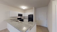 This single-story residence at 75 Northbrook Circle in Fairview Heights, Illinois, offers a well-designed, open layout with consistent finishes throughout. The kitchen serves as a central highlight, featuring granite countertops that extend across a peninsula breakfast bar, white shaker-style cabinetry reaching toward the ceiling, and a full suite of black appliances including a range, over-the-range microwave, and refrigerator. Wood-look hard flooring in the kitchen transitions into the adjoining living area, where plush neutral carpeting extends across a generously sized, open-concept space. A ceiling fan with light fixture overhead and a sliding glass door fitted with vertical blinds bring both airflow and natural light into the living area. The bedrooms are carpeted and well-proportioned, each equipped with ceiling fans and windows with blinds. Two bathrooms mirror the kitchen's design aesthetic, with granite-topped white vanities, wood-look flooring, and well-lit mirrors. A dedicated laundry area accommodates a stacked Whirlpool washer and dryer unit. Throughout, the space maintains a clean, neutral palette with white walls and trim, creating a bright and cohesive atmosphere.