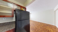 This single-floor residence offers a practical and well-designed layout that makes efficient use of its space. The living area is immediately welcoming, showcasing rich, patterned parquet hardwood flooring throughout that adds warmth and character to the interior. Neutral, light-toned walls keep the space feeling open and bright, complemented by a window that allows natural light to filter in.

The kitchen is a standout feature, outfitted with deep cherry-toned wood cabinetry accented by glass-panel upper cabinet doors and sleek bar-pull hardware. Dark granite countertops provide a bold contrast against the cabinetry, while black appliances — including a gas range and a full-size refrigerator — complete the cohesive, modern aesthetic. The kitchen flows openly toward the living area, creating a connected and sociable layout. Light tile flooring in the kitchen distinguishes it from the parquet in the adjoining spaces.

The bathroom is clean and classically styled, featuring white subway tile wainscoting that lines the lower walls, lending a timeless and tidy appearance. A window provides ventilation and a touch of natural light to this functional space.

Overall, the residence presents a cohesive design with consistent material choices, offering a comfortable and practical living environment with notable kitchen detailing and warm flooring throughout.