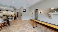 This inviting game room is designed for recreation and social enjoyment, offering a thoughtfully arranged layout across a single floor. The space is finished with warm-toned stone tile flooring set in a varied pattern, complemented by crown molding and neutral-painted walls that lend a polished, cohesive feel throughout.

At the heart of the room sits a full-size pool table, accompanied by a wall-mounted cue rack and a pair of classic gumball machines, adding a fun, nostalgic touch. A shuffleboard table provides additional entertainment, while a dining table with upholstered chairs offers a comfortable spot for casual seating or tabletop games.

A wall-mounted flat-screen television is positioned for easy viewing from multiple areas of the room. Recessed lighting and wall sconces provide warm, layered illumination throughout. Elegant French doors with glass pane detailing connect the game room to an adjacent living area, allowing natural light to flow freely between spaces.

Large windows dressed with decorative valances and horizontal blinds line one wall, offering generous views of a sparkling outdoor pool surrounded by lush greenery. The bright, airy atmosphere created by this natural light enhances the room's welcoming ambiance, making it an ideal gathering space for entertainment and leisure.