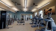 Located at 175 Harbor Bridge Ln in Mount Pleasant, South Carolina, this fitness center offers a thoughtfully designed single-floor layout built to accommodate a comprehensive workout experience. The space features a generous open floor plan with teal-accented walls and neutral carpeted flooring, creating a clean and energizing atmosphere.

The cardio section along the right side of the room is lined with modern treadmills and elliptical machines, positioned near a row of arched windows that allow abundant natural light to filter in. Multiple ceiling fans are evenly distributed throughout, promoting airflow and comfort during workouts. Overhead linear lighting supplements the natural light, ensuring the entire space is well-illuminated.

The strength training area occupies the remaining floor space and includes a variety of cable machines, weight benches, and a dumbbell rack. Floor-to-ceiling mirrors along one wall visually expand the space while allowing users to monitor their form. A separate glass-enclosed room at the rear appears to offer additional training or stretching space. Overall, the facility is well-maintained and functionally organized for diverse fitness needs.