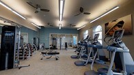 Located at 175 Harbor Bridge Ln in Mount Pleasant, South Carolina, this fitness center offers a thoughtfully designed single-floor layout built to accommodate a comprehensive workout experience. The space features a generous open floor plan with teal-accented walls and neutral carpeted flooring, creating a clean and energizing atmosphere.

The cardio section along the right side of the room is lined with modern treadmills and elliptical machines, positioned near a row of arched windows that allow abundant natural light to filter in. Multiple ceiling fans are evenly distributed throughout, promoting airflow and comfort during workouts. Overhead linear lighting supplements the natural light, ensuring the entire space is well-illuminated.

The strength training area occupies the remaining floor space and includes a variety of cable machines, weight benches, and a dumbbell rack. Floor-to-ceiling mirrors along one wall visually expand the space while allowing users to monitor their form. A separate glass-enclosed room at the rear appears to offer additional training or stretching space. Overall, the facility is well-maintained and functionally organized for diverse fitness needs.
