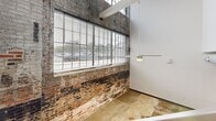 This two-floor loft in Burlington, NC showcases an industrial-chic aesthetic with exposed brick walls, soaring ceilings, and visible ductwork that celebrate the building's historic character. The kitchen features granite countertops, rich dark cabinetry, stainless steel appliances, and a large island with pendant lighting overhead. Polished concrete floors anchor the lower level, while warm hardwood flooring flows through the upper spaces. Oversized windows flood the interior with natural light, offering street-level views. Two full bathrooms include granite vanities, dark wood cabinetry, and tub-shower combinations. Multiple rooms provide flexible living arrangements throughout both floors.