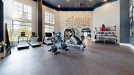 Located at 4129 Falcon Pkwy in Oakwood, Georgia, this impressive fitness center offers a spacious, single-floor layout thoughtfully designed to accommodate a full range of workout activities. The facility is divided into distinct zones, each tailored to a specific training discipline, yet unified by a cohesive and polished aesthetic.

The cardio area greets visitors with soaring double-height ceilings, large multi-pane windows that flood the space with natural light, and rubber flooring built to withstand heavy use. An assortment of treadmills, stationary bikes, rowing machines, and stair climbers are neatly arranged, while a dedicated rack keeps kettlebells and free weights organized and accessible.

The strength training zone features a deep navy blue accent wall adorned with a gallery of black-and-white athletic photography, lending an energetic and motivational atmosphere. Cable machines, functional trainers, and free-weight benches are strategically positioned throughout, with floor-to-ceiling mirrors lining the walls to maximize sightlines and create a sense of expansiveness. Oversized ceiling fans with elegant metallic finishes provide ample air circulation overhead.

A transition area with large mirror panels and refined tile flooring connects the fitness spaces to what appears to be an adjoining lobby or common area, featuring tall columns and grand architectural detailing. Throughout the facility, the design balances high performance with sophisticated style, making it a standout amenity.