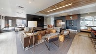 This contemporary amenity lounge is a thoughtfully designed social space that blends modern aesthetics with warm, inviting materials. The main lounge area is anchored by a striking dark-paneled fireplace wall mounted with a flat-screen television, set against a backdrop of large panoramic windows that frame sweeping urban skyline views and flood the space with natural light. A wood-planked ceiling detail adds warmth and architectural interest overhead, while large-format tile flooring grounds the room with a clean, polished finish. The lounge is furnished with an eclectic mix of tufted leather ottomans, sling chairs, plush sofas, and solid wood cube tables, all arranged atop a bold geometric-patterned area rug that defines the central gathering zone. A second, more casual seating area features comfortable sectionals and wood coffee tables positioned near floor-to-ceiling windows that open visually to an adjacent outdoor terrace. The fully equipped demonstration kitchen is outfitted with professional-grade stainless steel appliances including a gas range, dark wood cabinetry, open shelving, a white geometric tile backsplash, and an expansive concrete-topped island with vibrant orange bar stools — ideal for culinary events and social gatherings. Throughout the space, warm wood accents, industrial-inspired lighting, and curated décor create a cohesive, modern-urban atmosphere.