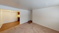 This single-floor residence offers a straightforward, well-organized layout designed for comfortable everyday living. The living area is generously sized, featuring neutral wall-to-wall carpeting that adds warmth underfoot, crisp white walls, and a broad window that invites natural light to fill the room. A baseboard heating system runs along the exterior wall, complemented by a wall-mounted air conditioning unit, ensuring year-round climate comfort. The space transitions smoothly into a connected area with wood-look flooring, leading to the kitchen. The kitchen is efficiently designed with rich, dark wood cabinetry providing ample storage, granite-style countertops, and black appliances including a full-size refrigerator, gas range, and over-the-range microwave. The wood flooring continues through this area, adding a cohesive and durable finish. A ceiling fan is visible in the adjoining area, contributing to the overall airflow and comfort of the space. The bathroom features a classic subway tile surround enclosing a full soaking tub with chrome fixtures, offering a clean and timeless aesthetic. Throughout the residence, the design prioritizes functionality with a neutral palette, making it an easy canvas to personalize.