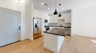 Located at 600 Arnold St NE in Atlanta's Old Fourth Ward neighborhood, this single-floor residence showcases a clean, modern design aesthetic carried consistently throughout the space. The kitchen serves as a centerpiece, featuring floor-to-ceiling white cabinetry, dark stone countertops, and a spacious central island illuminated by striking black pendant lights with gold interiors. A hexagonal tile backsplash adds visual texture, while stainless steel appliances — including a gas range, built-in microwave, and French door refrigerator — complete the contemporary kitchen setup. Light wood-tone flooring flows through the main living and kitchen areas, contributing to a bright, airy atmosphere enhanced by recessed lighting. The living area offers a generous, open layout with white walls and clean architectural lines. Bedrooms feature plush neutral carpeting, ceiling fans, and windows that bring in natural light and city views. The bathroom continues the modern theme with a long white vanity topped with dark stone countertops, pendant lighting, a large mirror, and large-format gray tile surrounding the bathtub. Gray panel-style doors and black hardware provide cohesive design details throughout the unit.