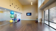 Located at 121 Towne St in Stamford, Connecticut, this fitness and wellness studio is a thoughtfully designed space built to accommodate a variety of exercise modalities. The studio features soaring double-height ceilings that create an open, airy atmosphere, complemented by rich dark hardwood flooring throughout. Large floor-to-ceiling windows along one wall flood the space with natural light and offer views of the surrounding urban streetscape.

The main exercise area provides generous open floor space, ideal for group fitness classes, yoga, stretching, or personal training sessions. A wall-mounted flat-screen television displaying a Fitness on Demand platform allows users to access guided workout programming at any time. An organized equipment rack along one wall holds a colorful assortment of stability balls, medicine balls, resistance bands, and exercise mats, while a dumbbell rack offers a range of free weights for strength training.

A frosted glass partition wall adds a modern architectural touch, visually separating a secondary workout area while maintaining a sense of openness. Additional amenities include a wet bar or refreshment station with a granite countertop, a stainless steel waste receptacle, and a glass-enclosed entryway with steel framing that contributes to the studio's contemporary aesthetic. The overall ambiance is clean, functional, and welcoming, suited for residents or members seeking a well-equipped fitness environment.