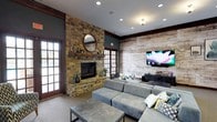 This inviting single-floor space blends rustic warmth with contemporary design, creating a welcoming atmosphere well-suited for both casual lounging and social gatherings. The focal point of the room is a striking floor-to-ceiling natural stone fireplace, complete with a richly stained wooden mantle adorned with decorative accents and a round mirror. Flanking the fireplace are classic French doors that allow natural light to filter into the space, enhancing the room's warm and open ambiance. Opposite the fireplace, a reclaimed wood accent wall adds texture and character, serving as a backdrop for a large wall-mounted flat-screen television and a sleek media console below. The seating area is anchored by a spacious gray sectional sofa accented with patterned throw pillows in teal and yellow, paired with a minimalist rectangular coffee table. Recessed lighting in the ceiling provides even, ambient illumination throughout, while dark wood crown molding adds a refined finishing touch. The overall design thoughtfully balances natural materials with modern furnishings, resulting in a space that feels both comfortable and polished.