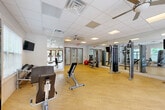 Located at 1321 Lovvorn Rd in Carrollton, Georgia, this dedicated fitness facility offers a thoughtfully designed, single-floor exercise space that accommodates a comprehensive range of workout options. The room features light hardwood-style flooring throughout, providing a clean and durable surface well-suited for fitness activities.

Natural light flows generously into the space through multiple large windows fitted with blinds, while glass-paned double doors on one end further enhance the bright, open atmosphere with views of the surrounding greenery. Ceiling fans are installed overhead to maintain airflow and comfort, complemented by a drop tile ceiling with recessed panel lighting for consistent illumination throughout.

Full-length mirrors line a significant portion of the walls, visually expanding the space and supporting proper form during workouts. The cardio zone features multiple treadmills and elliptical machines, while the strength training area includes a multi-function cable machine, adjustable benches, a dumbbell rack, medicine balls, and stability balls. A wall-mounted flat-screen television is positioned for convenient viewing during exercise.

An additional adjoining room or alcove is accessible from the main floor, adding functional utility to the facility. The overall design is clean, organized, and purposeful, making this fitness center a highly functional space for a well-rounded exercise routine.