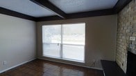 This single-story space presents a straightforward and functional interior defined by a few notable architectural details. The room features a textured popcorn ceiling accented by dark-painted exposed beams, creating a visual contrast against the neutral beige walls. Dark trim work frames the room's perimeter, adding definition to the overall design.

A prominent large picture window serves as the focal point of the room, fitted with horizontal blinds that allow for adjustable natural light. The window offers a view of greenery outside, connecting the interior to the outdoor surroundings. The generous size of the window ensures the space is well-lit throughout the day.

The flooring consists of square tile in earthy, warm tones, providing a durable and easy-to-maintain surface. Baseboards run along the walls, and a standard electrical outlet is visible near the window. The room's layout is open and unencumbered, offering flexibility for various uses. The combination of the textured ceiling, dark trim, and tiled flooring gives the space a casual, utilitarian character suited to practical everyday living.