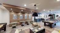 This single-story communal space in Dallas showcases a contemporary design that blends comfort and functionality. The interior is anchored by a striking wood-paneled accent wall adorned with a series of framed botanical artwork, adding a natural, earthy warmth to the modern aesthetic. Recessed lighting runs throughout the ceiling, supplemented by a bold, oversized black pendant light that serves as a dramatic focal point above the central lounge area.

The space is thoughtfully divided into distinct zones for varied uses. A casual seating area features a mix of lounge chairs and a low coffee table stocked with books and games, including a chess set, inviting relaxed gatherings. Nearby, small café-style tables paired with wire-frame chairs line the wood-paneled wall, offering intimate spots for work or conversation. A pool table positioned toward the far end of the room adds a recreational element, while a reception or service counter with teal accents is visible in the background.

Light wood flooring ties the space together, complementing the neutral color palette punctuated by subtle pops of color in the furnishings and rug. The open layout encourages flow and interaction throughout.