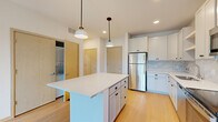 This contemporary single-floor residence showcases a thoughtfully designed open-concept layout with a bright, neutral color palette and light hardwood flooring that runs throughout the space. The kitchen serves as a central hub, featuring white shaker-style cabinetry with black hardware, a marble-look tile backsplash, and a full suite of stainless steel appliances including a refrigerator, range, microwave, and dishwasher. A generously sized center island with a clean white countertop and pendant lighting above offers additional prep space and casual seating. The kitchen flows seamlessly into the living area, which benefits from abundant natural light and direct access to a private balcony through sliding glass doors, offering pleasant outdoor views of surrounding greenery and neighboring buildings. The bedrooms are spacious and bright, with large windows, light hardwood floors, and ceiling fans for comfort. One bedroom also features balcony access via a sliding door. The bathroom is well-appointed with a wide vanity, white cabinetry with black hardware, a large mirror, and stylish globe sconce lighting. An in-unit laundry area is also visible, adding everyday convenience. The overall ambiance is clean, modern, and move-in ready.