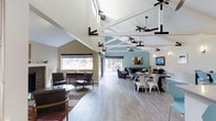 This single-story clubhouse offers a welcoming and well-appointed communal environment defined by its striking architectural character. The most notable feature is the dramatic vaulted ceiling with exposed white structural trusses accented by bold black hardware and multiple ceiling fans, creating an airy, open atmosphere throughout the main gathering area.

The space is organized into distinct functional zones that flow seamlessly into one another. A casual lounge area near the windows features mid-century modern seating arrangements on a patterned area rug, ideal for relaxed conversation. Adjacent to this is a dining area furnished with white chairs and dark tables, providing a versatile setting for meals or group activities.

Toward the rear of the space, a more intimate lounge area is anchored by a teal accent wall adorned with artwork and a wall-mounted television, complemented by contemporary sofas and additional seating. Large sliding glass doors and windows along the perimeter flood the interior with natural light and offer views of the surrounding greenery. Light-toned hardwood-style flooring runs throughout, tying the various zones together cohesively and contributing to the clubhouse's modern, inviting ambiance.