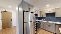 Located at 205 S Huntington Ave in Boston's Jamaica Plain neighborhood, this one-bedroom, one-bathroom unit offers a well-designed layout across a single floor. Light-toned hardwood flooring flows consistently throughout the space, creating a cohesive and clean aesthetic.

The kitchen is a standout feature, equipped with stainless steel appliances including a full-size refrigerator, gas range, and microwave. Gray shaker-style cabinetry with matte black hardware pairs with a striking blue mosaic tile backsplash and white quartz countertops. The open connection to the living area allows for flexible use of the space.

The living room is bright and airy, benefiting from multiple windows that frame views of surrounding greenery and invite generous natural light. The white walls and clean lines contribute to an open, modern feel.

The bedroom is a comfortable size and is served by multiple windows, continuing the theme of natural light. A dedicated closet with full-width wire shelving provides practical storage.

The bathroom features a warm wood-tone vanity with white quartz countertop, matte black fixtures, and a backlit mirror framed in black. A dark accent wall adds a modern, sophisticated contrast to the otherwise light and neutral palette of the unit.