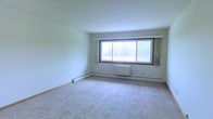 This single-floor space offers a clean, functional layout designed for comfortable everyday living. The main living area is notably spacious, featuring soft wall-to-wall carpeting that adds warmth underfoot and contributes to the room's calm, neutral aesthetic. Large, wood-framed windows span a generous portion of one wall, flooding the interior with natural light and offering pleasant views of the surrounding greenery and trees outside. The light-toned walls complement the bright atmosphere, creating an open and airy feel throughout. Practical baseboard heating runs along the perimeter of the room, ensuring consistent warmth, and a wall-mounted heating unit below the windows provides additional climate control. The overall design is straightforward and understated, with smooth ceilings and clean lines that lend the space a timeless, unfussy character. The single-floor layout makes for easy navigation and accessibility, while the abundance of natural light and connection to the outdoor landscape give the space an inviting and peaceful ambiance. Whether used as a primary living area or adapted to a variety of purposes, this space provides a solid, well-lit foundation.