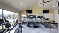 Located at 2223 East 151st Street in Carmel, Indiana, this fitness center offers a well-equipped, single-floor exercise facility designed to support an active lifestyle. The space is defined by its striking vaulted ceilings, which lend an open and airy atmosphere, while recessed lighting and a ceiling fan ensure the environment remains bright and comfortable during workouts. Large windows along one wall flood the space with natural light and provide views of the surrounding outdoor area, creating an inviting workout environment. Wall-to-wall mirrors along the back of the room enhance the sense of space and allow users to monitor their form during exercise. The facility is outfitted with a variety of cardio equipment, including multiple treadmills, elliptical machines, and a stationary recumbent bike, catering to a range of fitness preferences and routines. Multiple flat-screen televisions are mounted on the walls, offering entertainment options to keep members engaged during their sessions. The neutral color palette — featuring soft yellow accent walls and light-toned flooring — contributes to a clean, modern aesthetic throughout the space. Overall, this fitness center combines functional design with a welcoming ambiance, making it a practical and enjoyable destination for daily exercise.