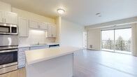 This well-designed single-floor residence offers a clean, contemporary aesthetic throughout, characterized by neutral tones, abundant natural light, and thoughtful finishes. The open-concept kitchen serves as a central hub, showcasing white shaker-style cabinetry, a full suite of stainless steel appliances including a refrigerator, range, microwave, and dishwasher, and a central island with a white countertop that provides additional workspace and casual dining potential. Light wood-tone flooring runs through the kitchen and living areas, adding warmth and continuity to the space. The living area is generously proportioned and features sliding glass doors that open directly to a private balcony, offering views of surrounding greenery and filling the interior with natural light. The bedrooms are carpeted for comfort and each benefit from large windows that frame pleasant outdoor scenery, including mature trees and open sky. The bathroom is finished with a wide white vanity, ample drawer and cabinet storage, brushed nickel hardware, and a walk-in shower. Throughout the residence, the design emphasizes clean lines, a light color palette, and practical layouts that create a comfortable and inviting living environment.