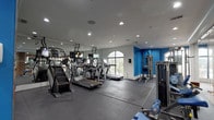 Located at 415 Armour Dr in Atlanta's vibrant 30324 zip code, this fitness center is a well-equipped, single-floor facility designed to support a comprehensive workout experience. The main exercise floor is finished with durable rubber matting and bathed in natural light streaming through multiple arched windows, complemented by recessed ceiling lighting throughout. The space features a robust selection of cardio equipment, including treadmills, ellipticals, and stair climbers, many equipped with individual display screens. Strength training is well represented with a cable crossover machine, a multi-functional cable station, a weight bench, and a dumbbell rack. Full-length mirrors along one wall enhance the sense of space and allow for form monitoring during workouts. A bold blue accent wall adds a dynamic visual element and houses a cork bulletin board and a wall-mounted water fountain station with seating, providing a convenient hydration and information area. An adjacent glass-partitioned space with hardwood flooring — visible from the main gym floor — appears to serve as a racquetball court or multipurpose activity room, adding versatility to the overall facility. The clean, bright, and energetic atmosphere makes this fitness center a functional and motivating environment for all fitness levels.