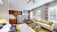 This single-floor unit offers a thoughtfully designed open-concept layout, where the living, sleeping, and dining areas flow seamlessly together. The galley-style kitchen is well-appointed with rich warm-toned cabinetry, granite countertops, a subway tile backsplash, and a full suite of stainless steel appliances, including a refrigerator, range, microwave, and dishwasher. The open living area is bathed in natural light from generously sized windows dressed with layered blinds and patterned drapes, creating a bright and airy atmosphere. A ceiling fan overhead adds both function and a casual elegance to the space. The sleeping area integrates naturally within the main room, complemented by an accent wall in a soft blue-gray tone that adds depth and visual interest. Hardwood-style flooring runs throughout, lending a cohesive and contemporary feel. The bathroom features large-format tile flooring, a granite-topped vanity with warm cabinetry, and a full tub-shower combination with white subway tile surround. Wall-mounted shelving provides additional storage and display opportunities throughout the unit.