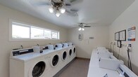 This single-floor communal laundry facility is designed for efficiency and comfort, offering residents a clean and functional space for their laundry needs. The room is lined with a full row of front-loading washing machines along one wall, paired with a corresponding lineup of dryers, providing ample capacity for multiple users simultaneously. The warm, neutral-toned walls and light-colored flooring contribute to a bright and welcoming atmosphere. A wide horizontal window along one wall invites natural light into the space, offering a pleasant view of the surrounding exterior. Two ceiling fans with light fixtures are evenly spaced throughout the room, ensuring consistent air circulation and comfortable temperatures during use. The textured ceiling adds a subtle architectural detail, while safety signage is thoughtfully posted on the walls. The layout is open and well-organized, allowing for easy movement between machines. Overall, this laundry facility is a practical, well-maintained shared amenity that prioritizes convenience and a comfortable user experience.