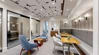 Located at 3500 Fairmount St in Dallas, TX, this thoughtfully designed single-floor business suite offers a refined blend of comfort and functionality across five distinct areas. The centerpiece of the space is a sophisticated lounge area distinguished by a striking geometric ceiling installation of layered white and wood panels, recessed lighting, and soft neutral tones. Plush blue accent chairs and warm wood side tables create an inviting setting for casual meetings or relaxation. Adjacent to the lounge is a café-style dining nook, where built-in banquette seating paired with natural wood tables and modern white chairs offers an ideal spot for informal gatherings. A dedicated beverage station with a coffee maker and counter space adds everyday convenience. A glass-enclosed private workstation provides a quiet, focused environment equipped with dual monitors, a height-adjustable desk, and an ergonomic chair — ideal for individual productivity. The conference room features a warm wood oval table, sleek rolling chairs, a wall-mounted display, a whiteboard, and natural light from tall windows framing lush outdoor greenery. Textured wall treatments, mood-enhancing sconce lighting, and cohesive neutral carpeting tie the entire suite together with a polished, professional ambiance.