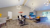 The Learning Center is a thoughtfully designed single-floor community space that seamlessly blends functionality with comfort. The main area is anchored by soaring vaulted ceilings and illuminated by striking Sputnik-style chandeliers that lend a modern, sophisticated feel. This open area is divided into distinct zones: a computer workstation section equipped with multiple desks and monitors for productivity, and a relaxed lounge area featuring a plush navy sectional sofa, colorful accent chairs, and a wall-mounted flat-screen television. Large sliding glass doors connect the lounge to an outdoor balcony, flooding the space with natural light and providing views of surrounding greenery.

Adjacent to the main area, a well-appointed kitchen includes full cabinetry, granite-style countertops, black appliances, and a peninsula bar with vibrant orange bar stools—ideal for casual gatherings or refreshment breaks. A separate dining area accommodates multiple round tables with upholstered seating, accented by floral centerpieces and framed artwork. An entry corridor with a sleek metal railing welcomes visitors and provides visual connectivity throughout. Patterned carpet tiles flow consistently across all areas, unifying the space with a clean, contemporary aesthetic.