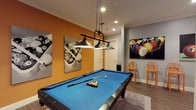 The Bullard Room is a stylishly designed billiards lounge that serves as a dedicated space for recreation and social gathering. At the heart of the room sits a full-size pool table with a striking blue felt surface, positioned beneath an industrial-style pendant light fixture with three wide-shade lamps suspended by chains — providing focused, atmospheric illumination over the playing surface. The flooring is a warm wood-tone laminate that grounds the space, while crown molding and clean white baseboards add a polished architectural touch. The room features a bold two-tone color scheme, with one wall painted in a vivid orange-amber hue that creates energy and contrast against the surrounding cool grey walls. Billiards-themed large-format canvas prints are displayed throughout, reinforcing the room's dedicated sporting character and adding visual interest at every angle. A wall-mounted cue rack keeps equipment neatly organized and accessible. A set of orange metal bar-height stools lines one wall, offering comfortable seating for spectators. Recessed ceiling lighting supplements the pendant fixture, ensuring the entire room is well-lit. The space is compact yet purposefully arranged, with clear circulation paths and doors providing easy access. Overall, the Bullard Room delivers a cohesive, game-focused environment ideal for casual play and social interaction.