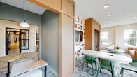 Located at 1630 Lewis Landing Ave in Wilmington, NC, this single-floor amenity space delivers a thoughtfully curated interior that balances bold design with functional versatility. The layout is defined by striking warm-toned architectural millwork frames that divide the space into distinct zones while maintaining an open, connected feel throughout.

The main lounge area features layered seating arrangements — including curved accent chairs, ottomans, and low coffee tables — set atop richly patterned area rugs that anchor the space. A standout wall displays a vibrant, abstract geometric wallcovering in terracotta, navy, and cream tones, serving as a bold focal point. Adjacent lounge nooks offer more intimate settings, with soft neutral upholstered chairs positioned near large windows that invite natural light and provide views of the surrounding greenery.

A dedicated kitchenette runs along one wall, outfitted with light wood cabinetry, open shelving, a sink, and countertop appliances — providing practical convenience for residents or guests. A private consultation or focus nook features floor-to-ceiling geometric wallcovering, a terrazzo-topped surface, and a modern wall sconce, creating an immersive and intimate atmosphere.

Black-framed glass partitions allow visual transparency between zones while defining separate areas for socializing or quiet retreat. Throughout the space, curated artwork, potted plants, and pendant lighting contribute to a polished, design-forward ambiance.
