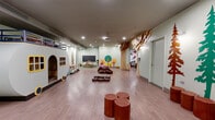 Located on Warren Street in Jersey City, NJ, this thoughtfully designed children's play space offers a richly immersive, nature-inspired environment spread across a single open floor. The space is anchored by a standout full-scale camper playhouse, complete with a lofted upper level and a miniature kitchen, serving as a central focal point for imaginative play. Smooth hardwood-style flooring extends throughout, providing a clean and safe surface for children to move and play freely.

The woodland theme is carried consistently through the decor: hand-painted tree murals in green and amber tones adorn the walls, while laser-cut plywood tree sculptures and a canopy archway add striking three-dimensional character. A nature-print mural wall featuring illustrated birds, insects, and animals creates a vivid, educational backdrop in one section of the space. A cozy teepee nestled among decorative evergreen trees offers a quiet retreat area, and tree-stump stools are scattered throughout for casual seating.

Recessed ceiling lighting illuminates the interior evenly, complemented by a ceiling-mounted projector for multimedia use. A chalkboard wall and small activity tables support learning and creative engagement. Brown bean bag chairs provide relaxed, flexible seating. The space also includes a separate room accessible via a private door, ideal for additional programming or staff use.