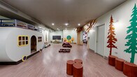 Located on Warren Street in Jersey City, NJ, this thoughtfully designed children's play space offers a richly immersive, nature-inspired environment spread across a single open floor. The space is anchored by a standout full-scale camper playhouse, complete with a lofted upper level and a miniature kitchen, serving as a central focal point for imaginative play. Smooth hardwood-style flooring extends throughout, providing a clean and safe surface for children to move and play freely.

The woodland theme is carried consistently through the decor: hand-painted tree murals in green and amber tones adorn the walls, while laser-cut plywood tree sculptures and a canopy archway add striking three-dimensional character. A nature-print mural wall featuring illustrated birds, insects, and animals creates a vivid, educational backdrop in one section of the space. A cozy teepee nestled among decorative evergreen trees offers a quiet retreat area, and tree-stump stools are scattered throughout for casual seating.

Recessed ceiling lighting illuminates the interior evenly, complemented by a ceiling-mounted projector for multimedia use. A chalkboard wall and small activity tables support learning and creative engagement. Brown bean bag chairs provide relaxed, flexible seating. The space also includes a separate room accessible via a private door, ideal for additional programming or staff use.