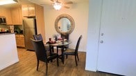 This single-floor residential space offers a comfortable and functional layout designed for everyday living. The dining area serves as a welcoming focal point, highlighted by rich hardwood-style flooring that adds warmth and character to the room. A round glass-top dining table paired with sleek black leather chairs provides an intimate setting for meals, while a decorative round mirror with an ornate metallic frame adorns the wall, adding a touch of elegance to the space. A ceiling fan overhead ensures year-round comfort and contributes to the room's casual, livable ambiance. Adjacent to the dining area, the kitchen is equipped with stainless steel appliances, including a full-size refrigerator, and features warm wood-toned cabinetry that complements the overall color palette of the home. The seamless flow between the kitchen and dining area creates an open, connected feel that is both practical and inviting. The neutral wall tones throughout provide a versatile backdrop, making the space easy to personalize. Overall, this home combines functional design with cozy, approachable details that create a comfortable living environment.