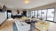 This contemporary single-floor amenity space is designed to foster comfort and social interaction through a thoughtfully open floor plan. The main lounge area is generously proportioned, featuring light hardwood flooring that flows throughout and anchors the space with a warm, inviting tone. Plush neutral-toned sectional sofas and accent chairs in bold yellow upholstery are arranged around a dark wood coffee table, creating a versatile seating area ideal for relaxing or gathering.

The kitchen and bar area is a standout feature, outfitted with dark cabinetry, a chevron-patterned tile backsplash, and a long dark-topped island with seating for multiple guests. A stainless steel refrigerator, microwave, and coffee station complete the functional setup, while pendant lighting overhead adds a refined, modern touch. A vibrant abstract artwork adds a pop of color against the neutral wall.

A secondary lounge nook at the far end of the space is bathed in natural light from a wraparound arrangement of large windows with adjustable striped roller shades, offering sweeping cityscape views. Teal leather accent chairs and a compact side table furnish this bright corner retreat.

Large black-framed sliding glass doors along one wall open directly to an outdoor terrace, blurring the line between indoor and outdoor living. A wall-mounted television and additional dining seating round out the amenities of this polished, urban communal space.