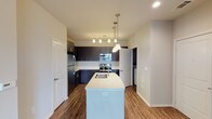 This single-floor, one-bedroom, one-bathroom unit offers a thoughtfully designed layout with contemporary finishes throughout. The kitchen serves as a central highlight, featuring dark espresso cabinetry that extends to the ceiling, providing ample storage space. Light-toned quartz countertops contrast beautifully against the dark cabinets, while a central island with a deep double-basin stainless steel sink adds both functionality and a natural gathering point. Stainless steel appliances, including a refrigerator, electric range, and over-the-range microwave, complete the kitchen. Pendant lighting above the island and a track light fixture illuminate the space effectively. Wood-style flooring runs throughout the main living areas, adding warmth and continuity. A dedicated laundry area is conveniently tucked behind a door adjacent to the kitchen. The bedroom offers plush carpeting, a ceiling fan, and direct access to the main living space. The bathroom features wood-style flooring, a tub/shower combination with floor-to-ceiling tile surround in neutral tones, and a vanity with dark cabinetry. The overall aesthetic is clean and modern, with neutral wall tones tying each space together cohesively.